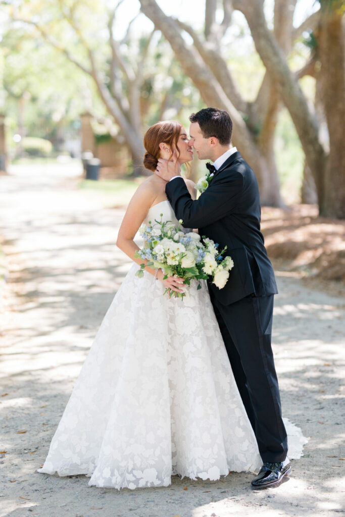 Bride in floral gown, groom kisses forehead outdoors.