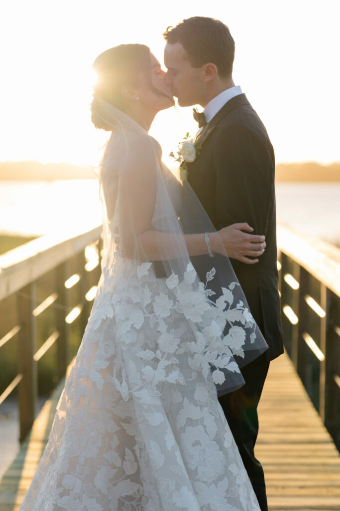 Bride and groom kiss on sunlit bridge at sunset.