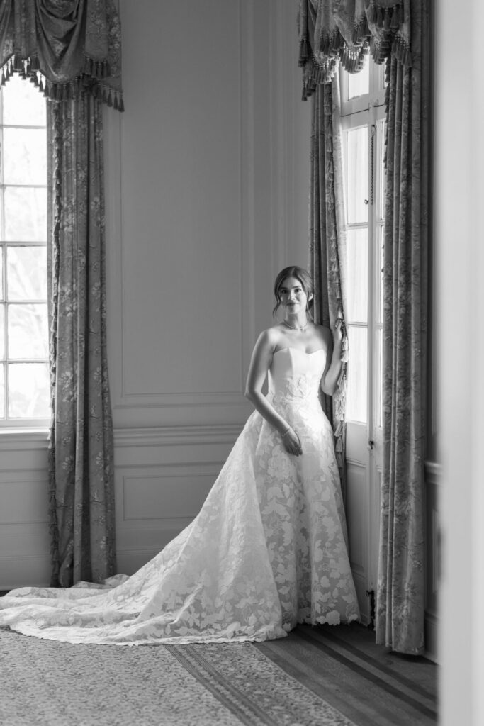 Bride in lace dress by window, black and white.