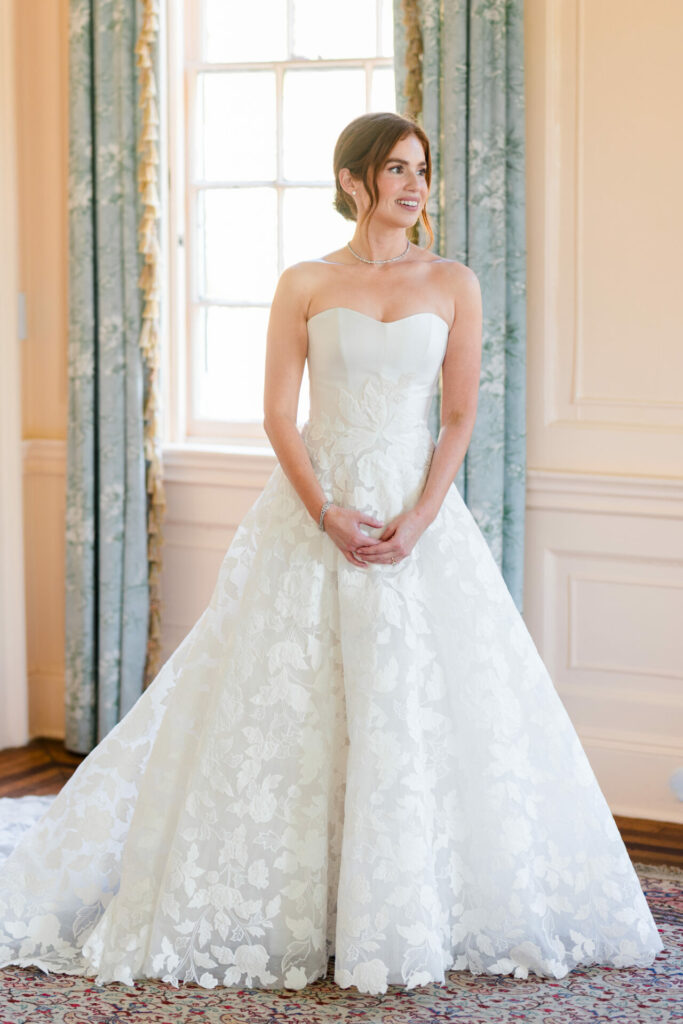 Smiling woman in floral wedding dress by window.