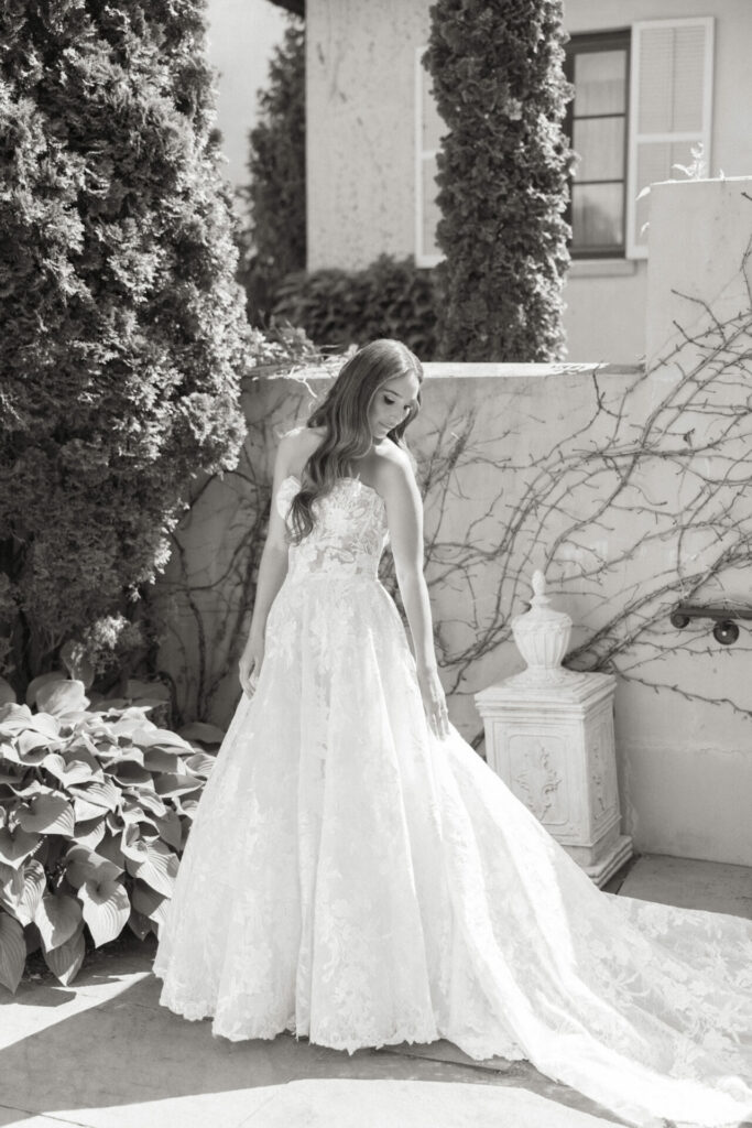 Bride in lace gown near sunlit, vine-covered wall.