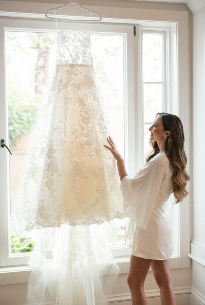 Woman in white robe admires a sunlit bridal gown.