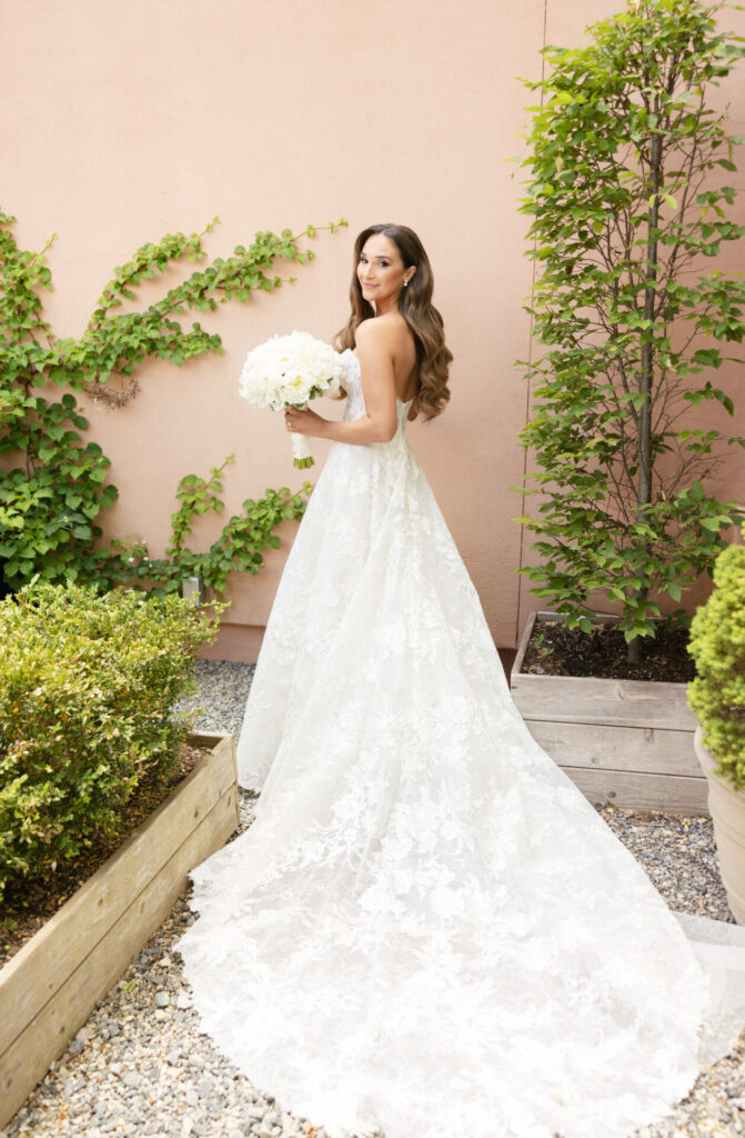 Bride in white lace gown outdoors, bouquet in hand.