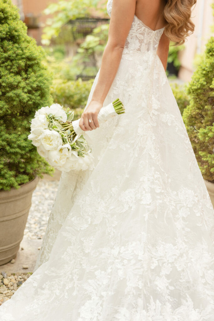 Bride in lace gown holds white bouquet outdoors.