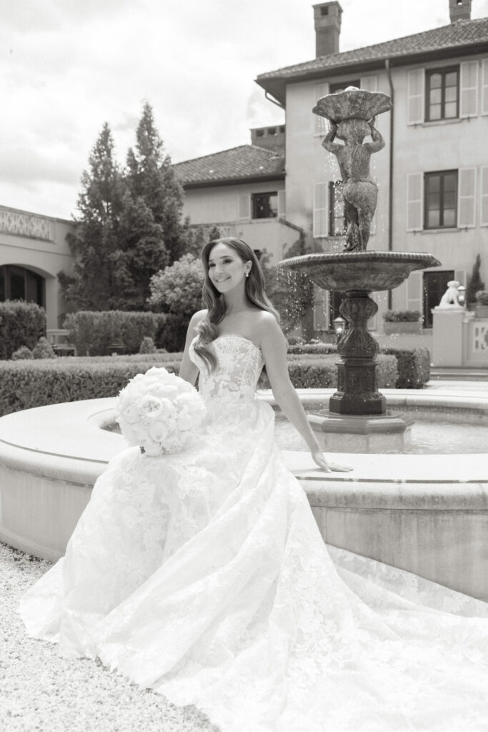 Bride in couture by fountain, holding flowers; elegant black-and-white wedding scene.