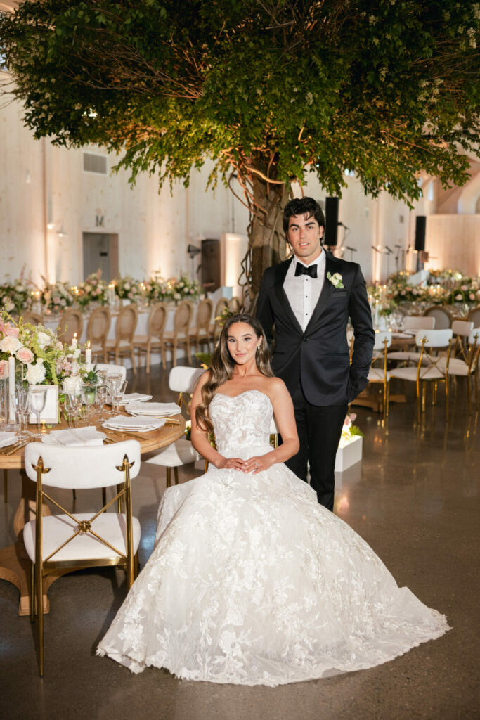Bride at elegant table, groom standing, floral indoor setting.
