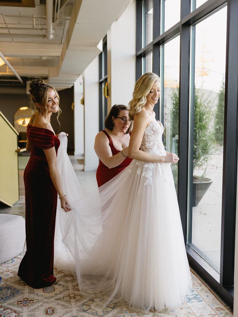 Bride in white, bridesmaids adjusting train by window.