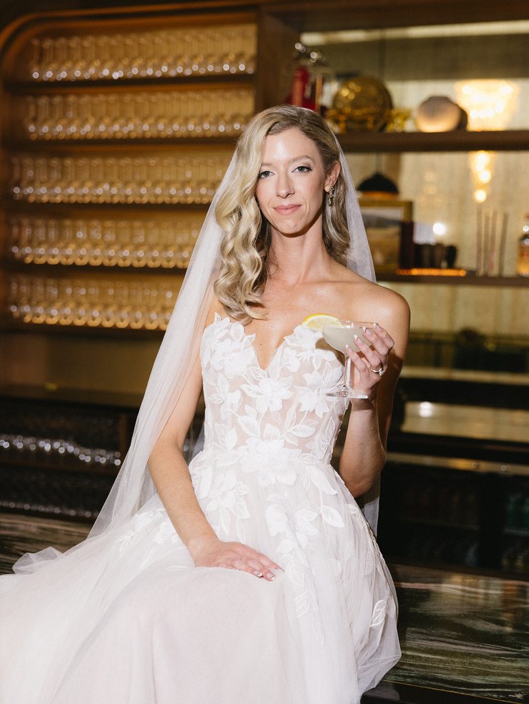 Smiling bride in couture gown holds cocktail at bar.