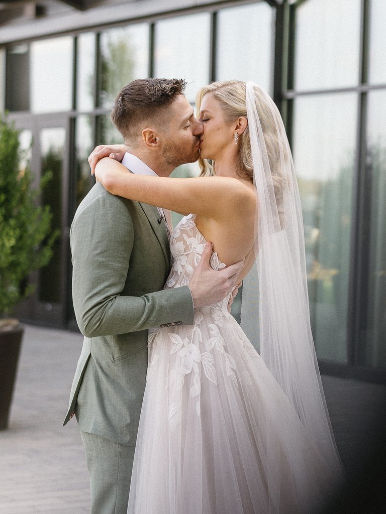 Bride kisses groom by glass building outdoors.