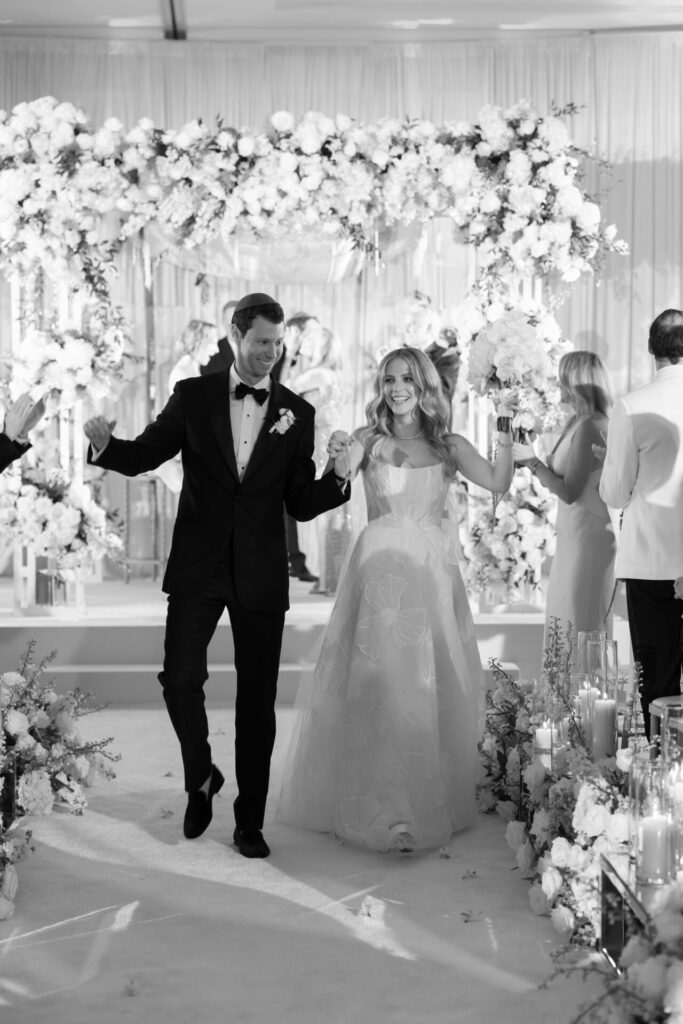 Smiling bride and groom walk aisle amid flowers.