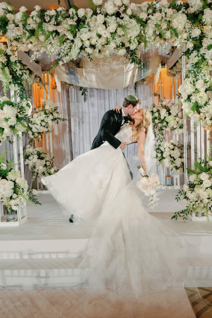 Groom dips, kisses bride under floral arch.