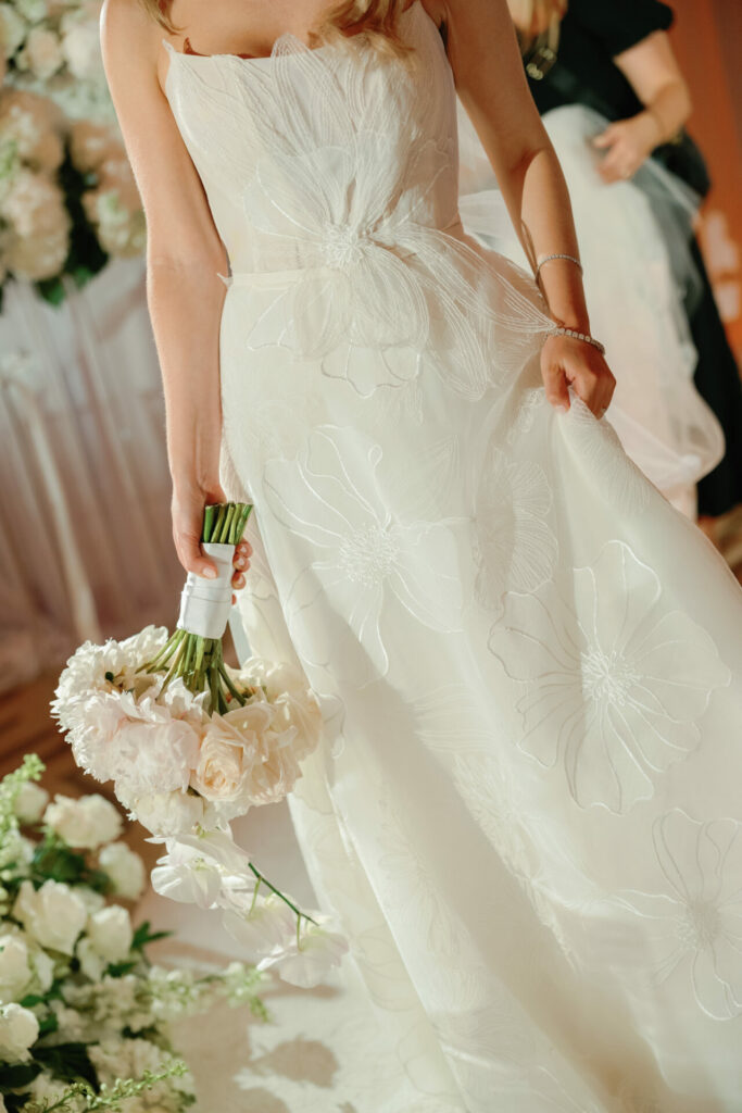 Bride in white floral dress with bouquet, elegant setting.