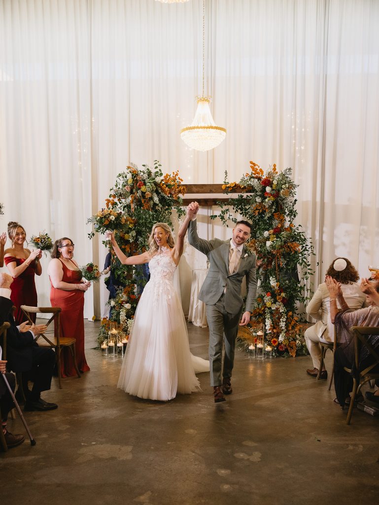Bride and groom walk aisle, smiling amid flowers.