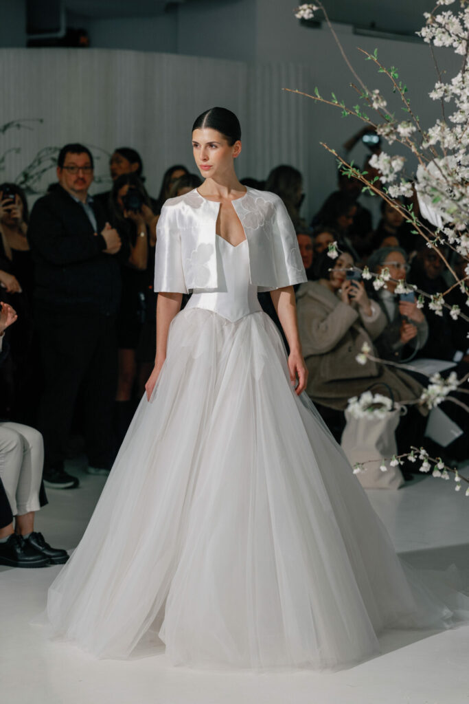Model in white dress by flowers on runway.