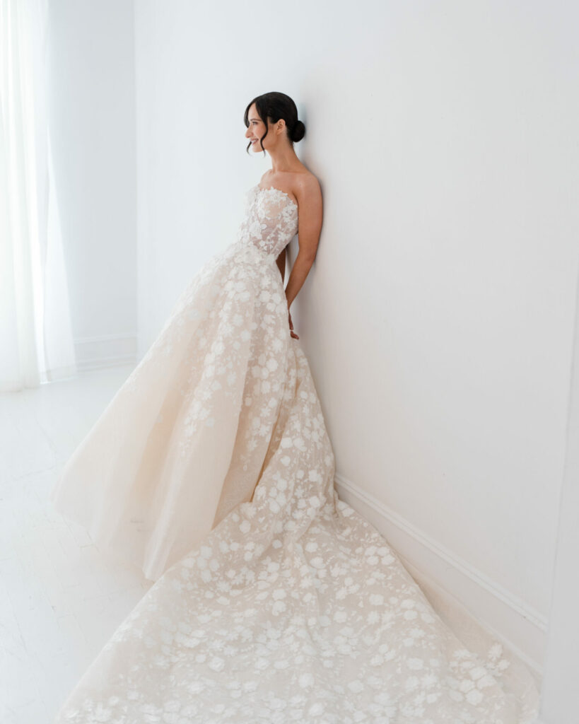 Bride in strapless lace gown leans against wall.