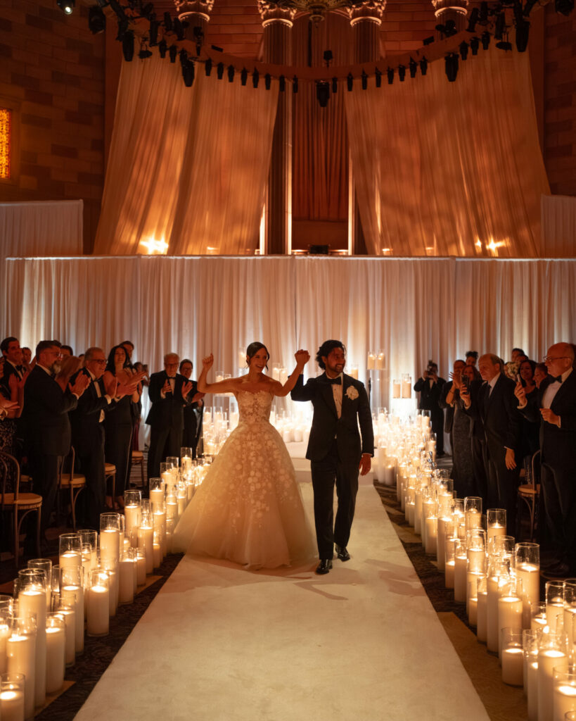 Bride and groom walk candlelit aisle, guests applaud.