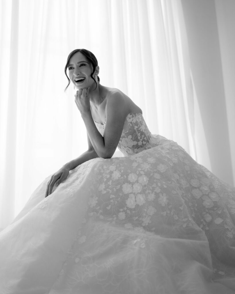 Smiling bride in lace dress by sheer curtains.