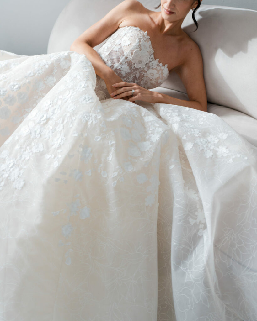 Bride in white floral gown reclines on sofa.