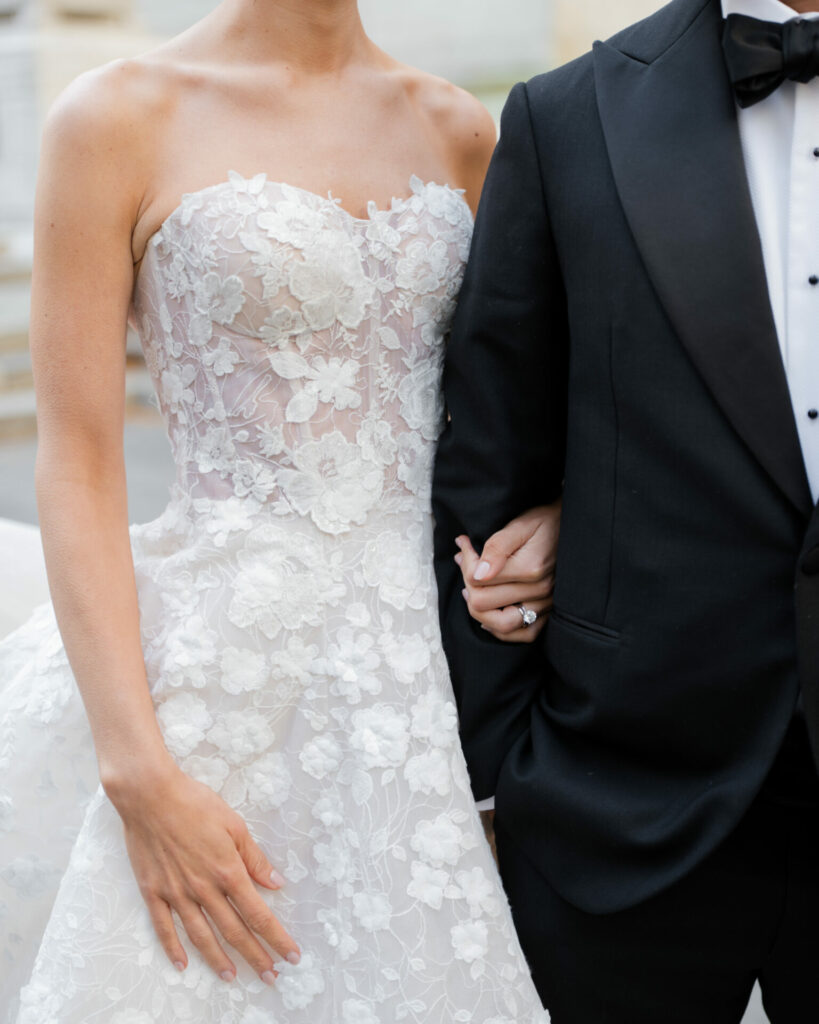 Bride in lace gown holds tuxedoed partner’s arm.