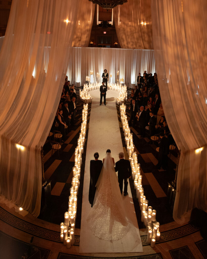 Bride walks candlelit aisle, sheer curtains glow.