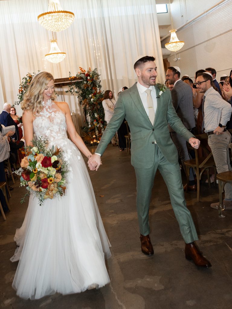 Bride and groom walk aisle, guests cheer, florals.