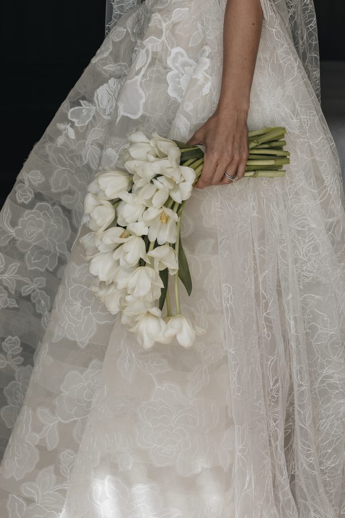 Bride in lace gown holding tulip bouquet, ring visible.