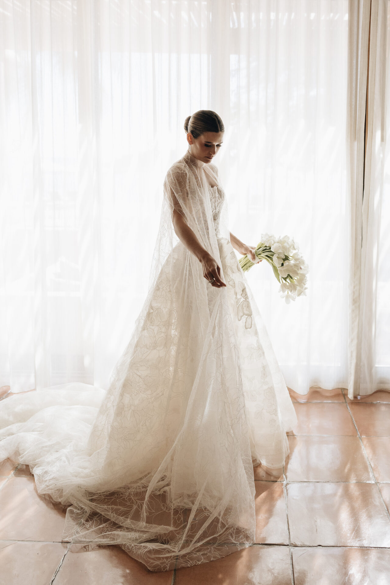 Bride in lace gown by sunlit sheer curtains.