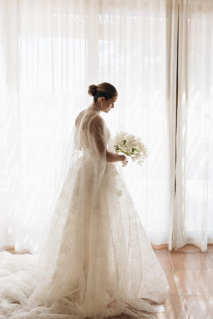 Bride in lace dress by sunlit sheer curtains.
