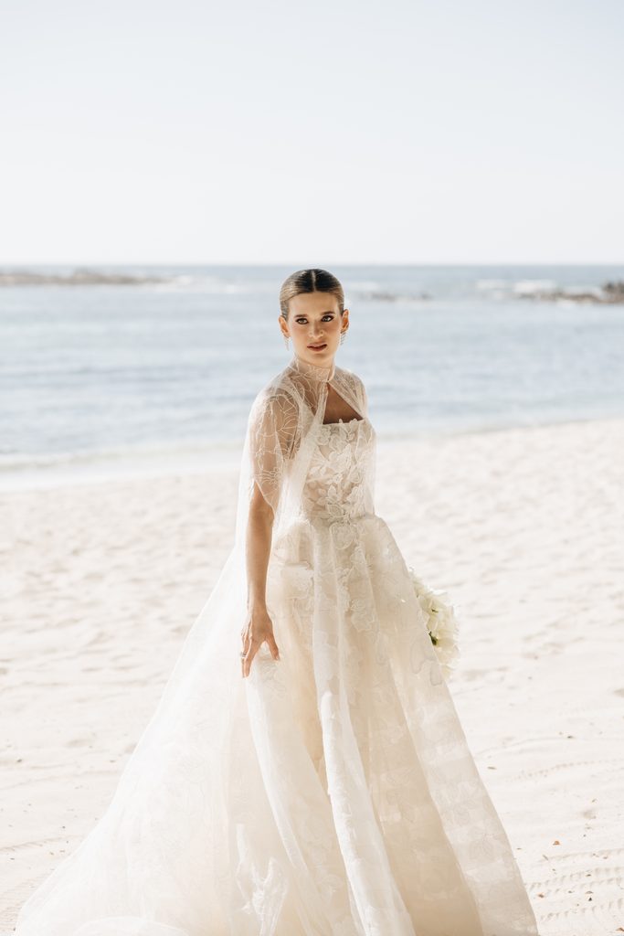 Bride in lace gown poses elegantly on beach.