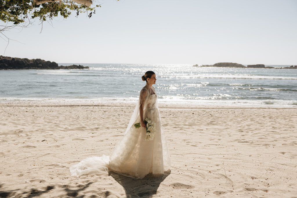 Bride in white dress walks alone on beach.