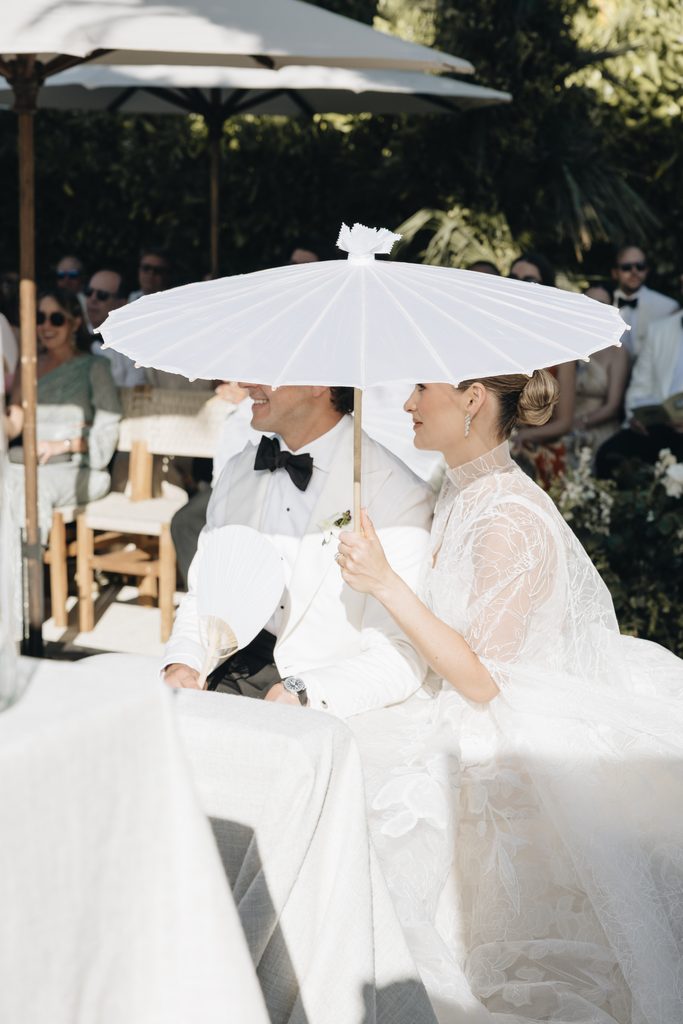 Bride and groom under parasol in white attire.