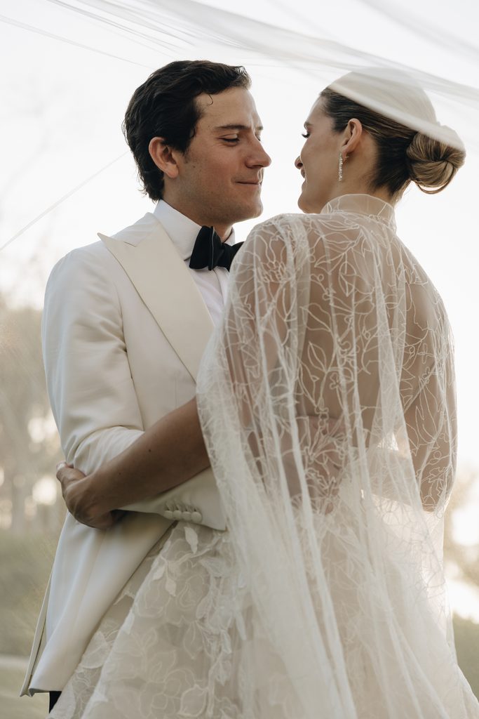 Bride and groom embrace outdoors under sheer embroidered veil.