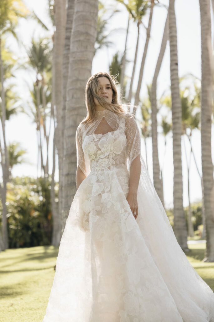 Bride in lace dress beneath sunlit palm trees.