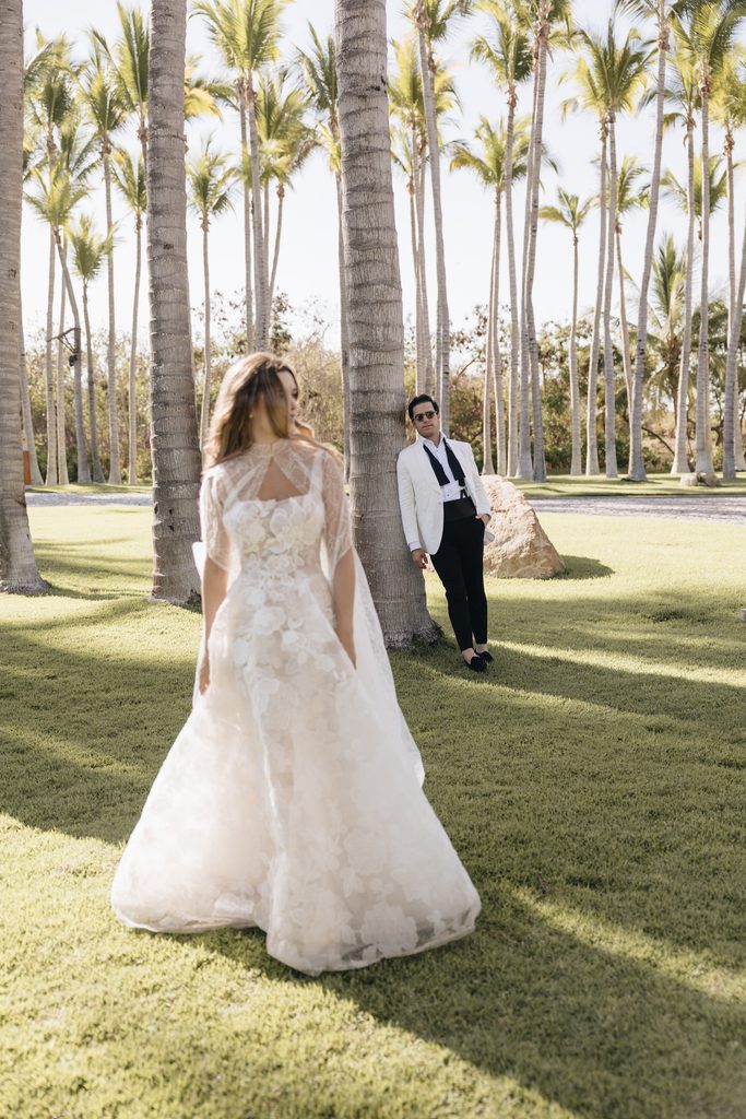 Bride in lace gown walks to tuxedoed partner outdoors.