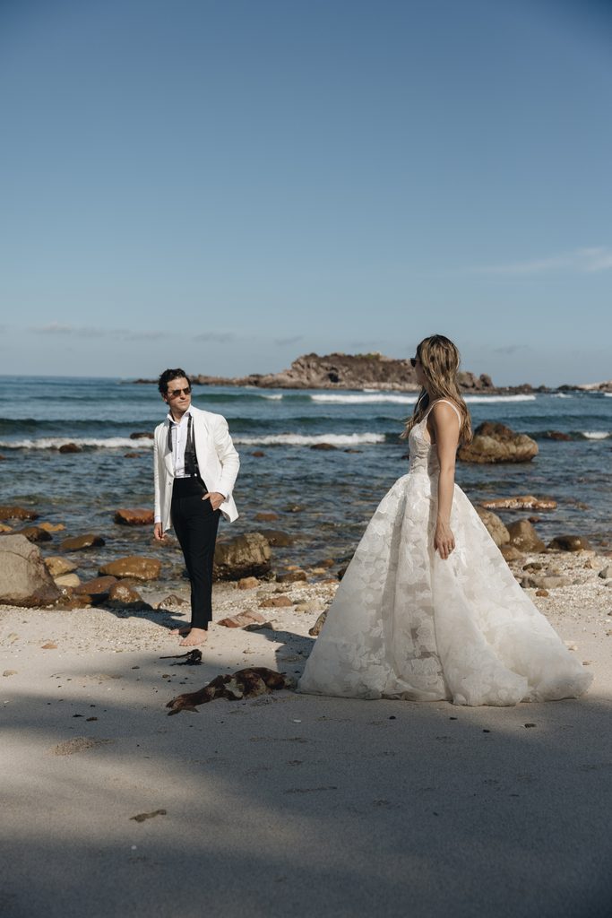 Bride and groom on rocky beach, clear sky.
