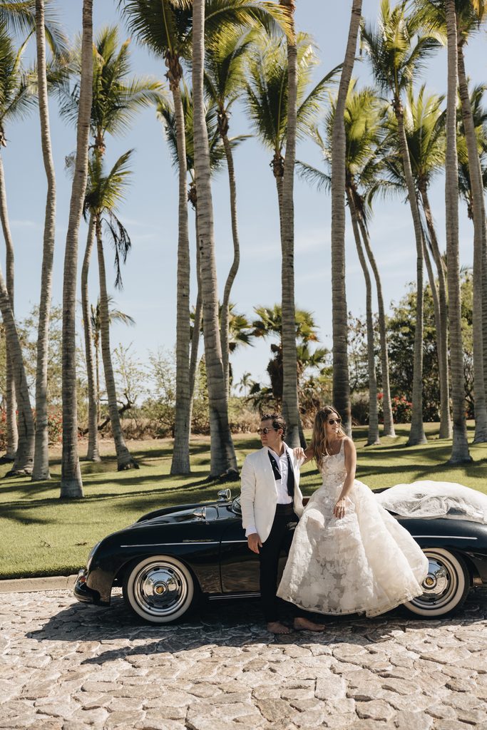 Bride and groom by vintage car under palm trees.