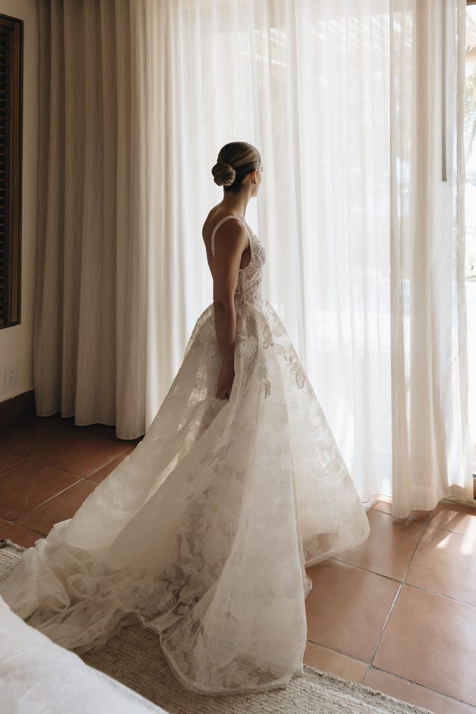 Bride in lace gown by sunlit window.