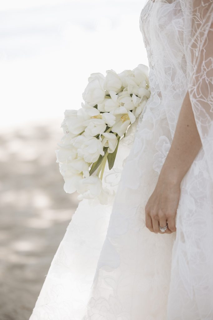 Bride in white lace, holding tulips outdoors.