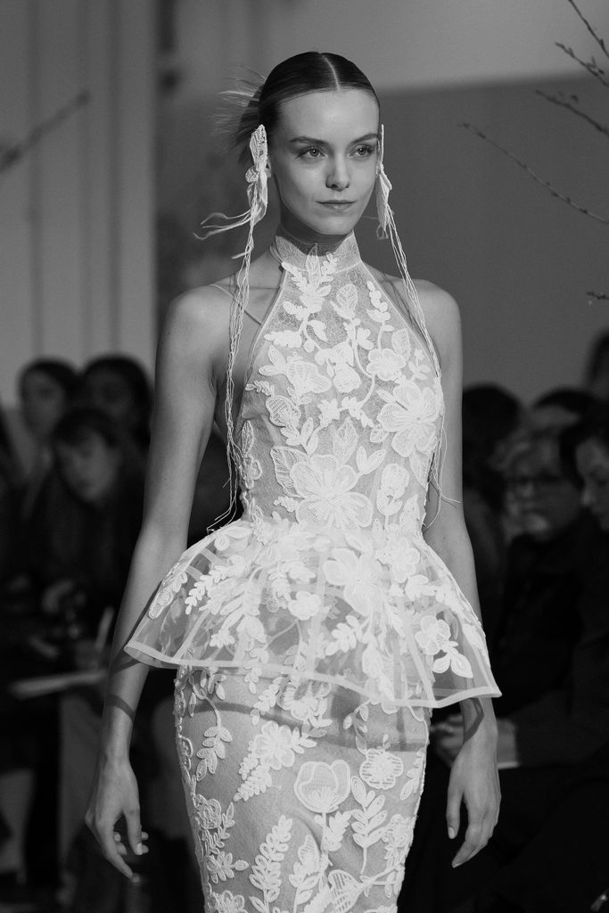 Model in lace dress on runway, black and white.