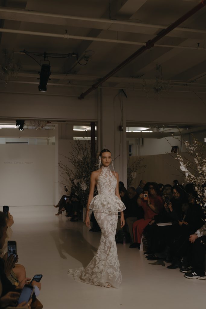Model in white lace gown on runway, watched.
